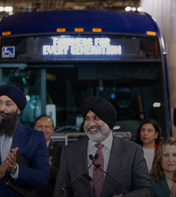 Smiling man speaking at podium in front of a bus with For Every Generation on display, surrounded by applauding audience.